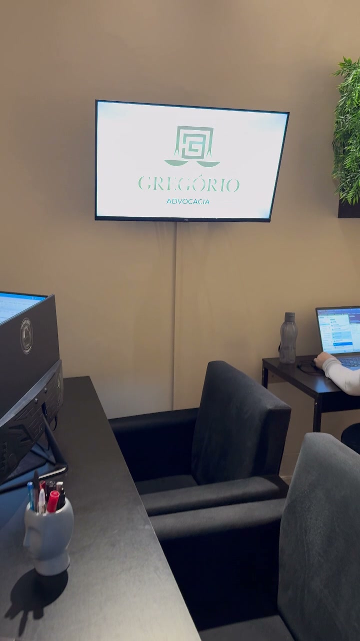 Sala de atendimento do escritório: mesa, duas poltronas e a logo Gregório Advocacia na televisão da parede.